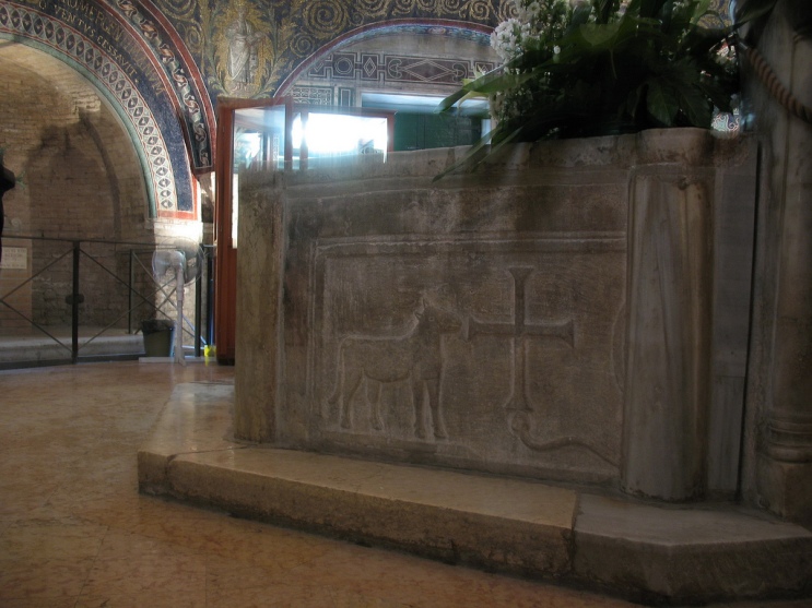 Λεπτομέρεια του εξωτερικού της κολυμβήθρας του ορθόδοξου βαπτιστηρίου της Ραβέννας (5ος αιώνας). Βλέπουμε ανάγλυφο έναν Αμνό με έναν Σταυρό.
